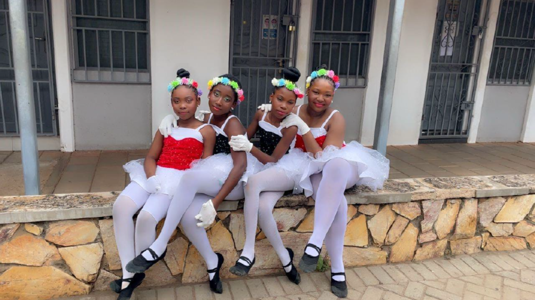 Young girl in ballet pose outdoors with other students and school buildings in background